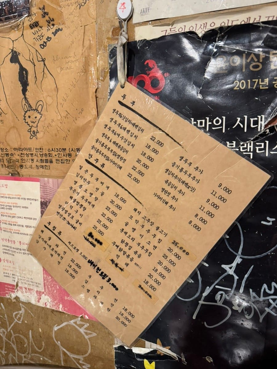 seoprano_가 남긴 사진입니다. 포스터가 겹겹이 붙은 벽에 걸린 두부 메뉴판과, 함께 나온 세 가지 기본 반찬을 담았습니다.