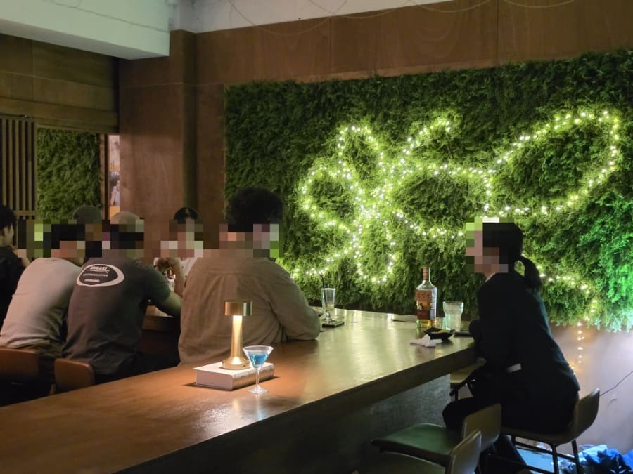 lovly1904가 담은 입구와 바 내부입니다. 우드톤 외벽에 自酌 간판이 은은하게 빛나고, 안으로 들어서면 이끼벽에 조명이 수놓인 바 테이블에 사람들이 나란히 앉아 있습니다.