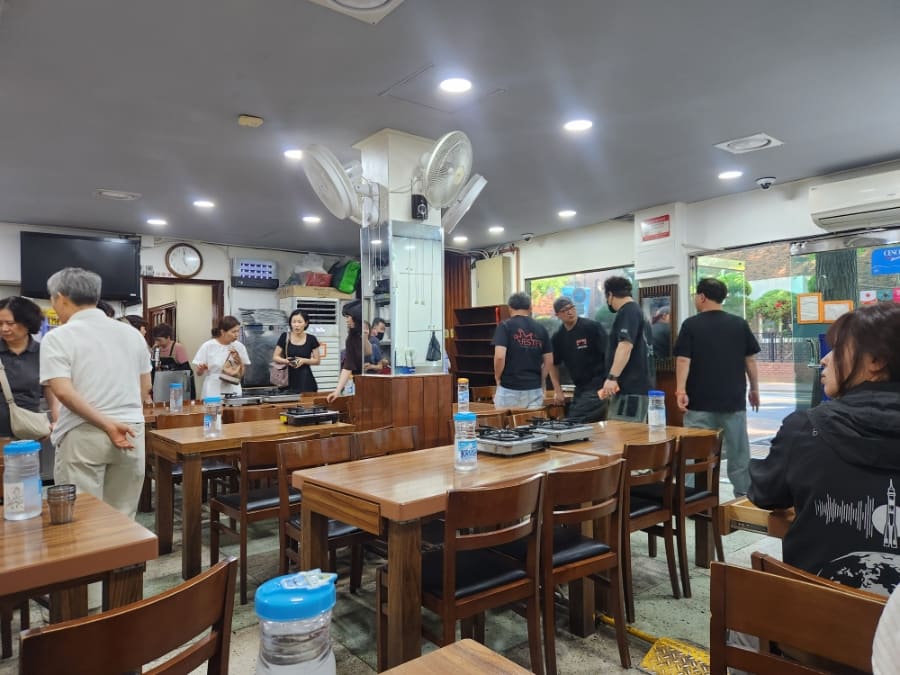 qozy는 평일 오후 영업을 열기 직전 가게 앞에 서 있는 사람들을 사진에 담았습니다. 나무 그늘 아래 붉은 벽돌 건물 앞으로 줄이 이어지고,