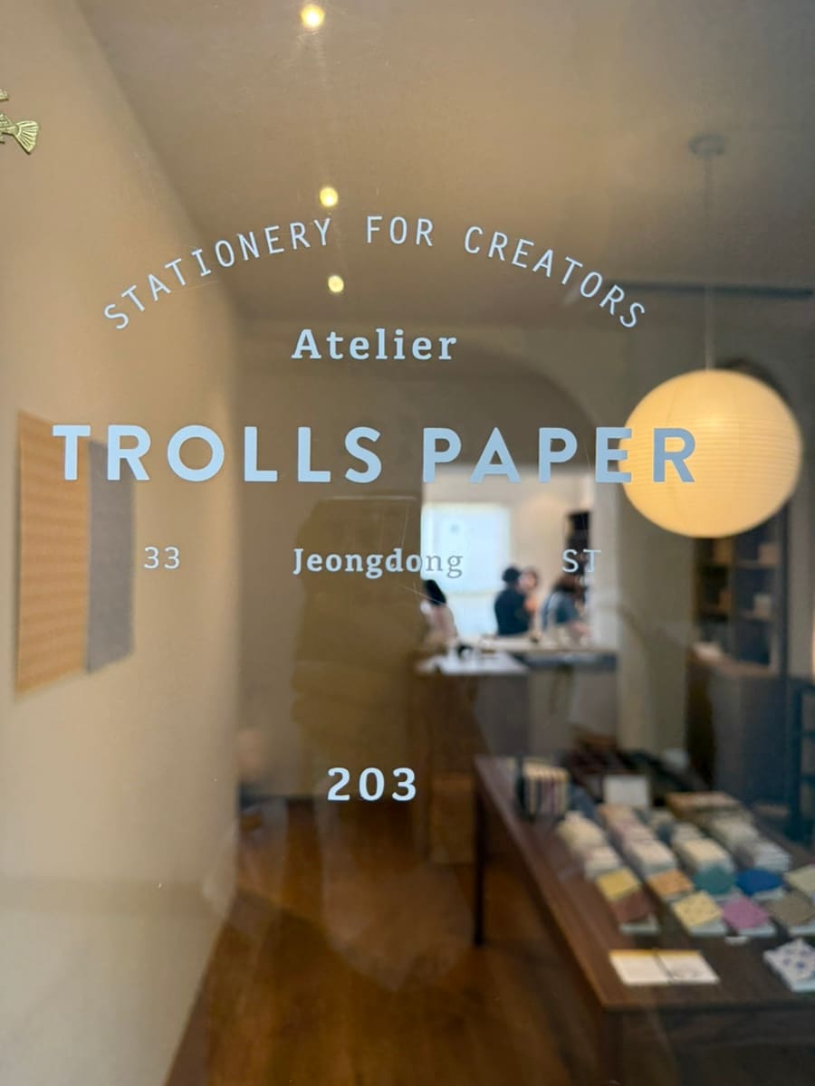 트롤스페이퍼의 입구 유리문에 보이는 간판입니다. 'STATIONERY FOR CREATORS Atelier TROLLS PAPER' 텍스트와 주소,