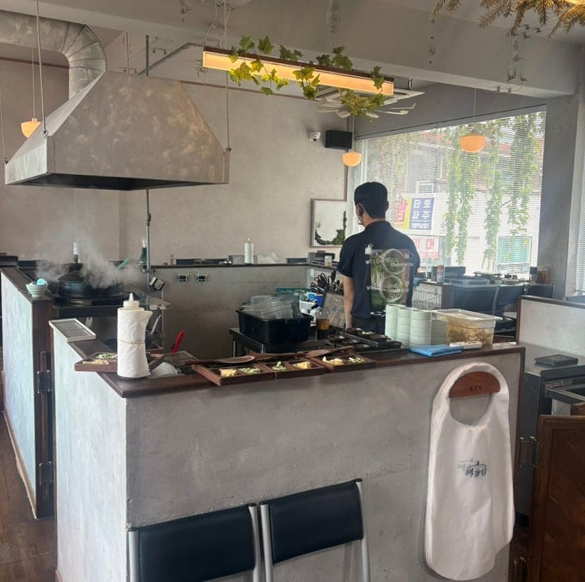 dailysuan이 9월 초, 두 번째 방문에서 남긴 사진입니다. 오픈 키친에서 연기가 오르고, 직원이 능숙하게 밑반찬을 준비합니다. '해율담'이라 새겨진 앞치마가 걸려 있고,