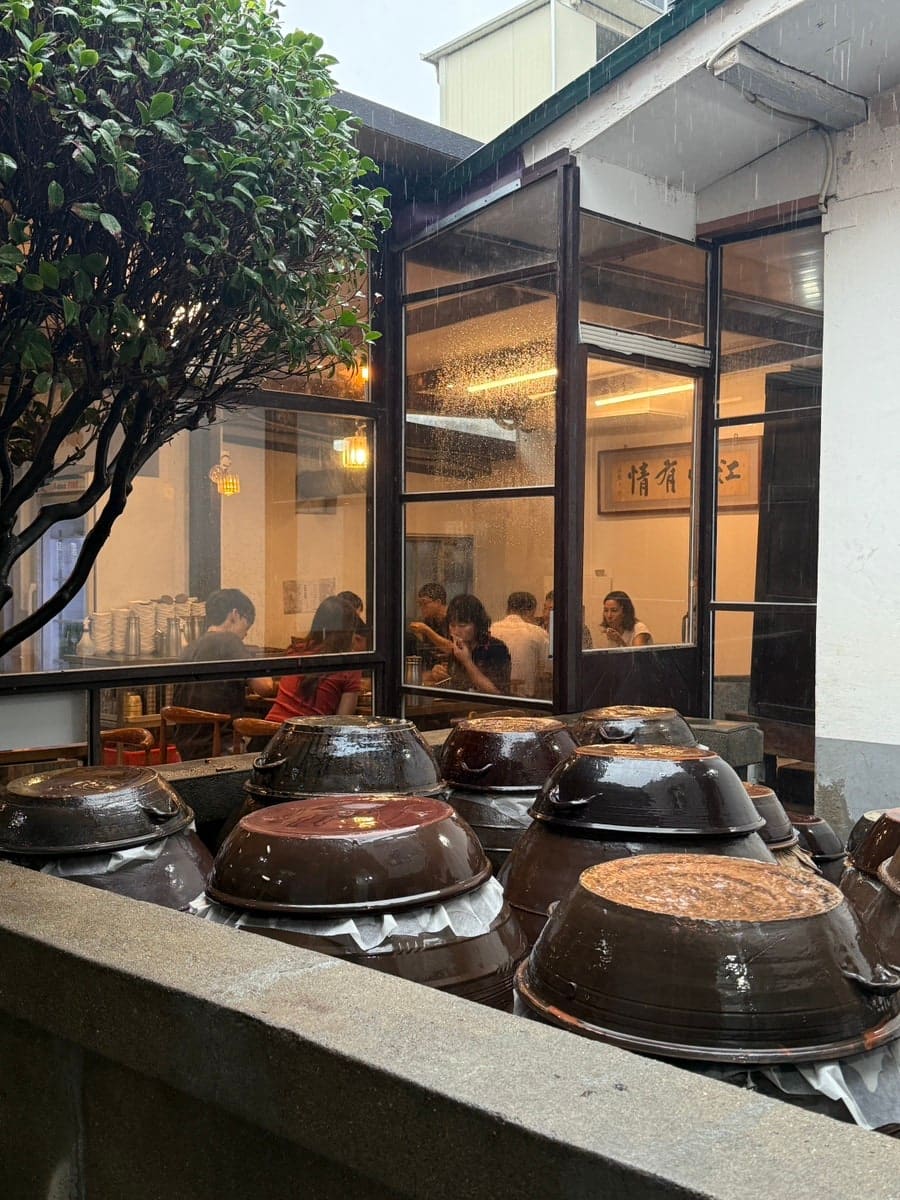 빗속에 놓인 장독 수십 개와, 통유리 안쪽에서 식사 중인 손님들이 보이는 노포 식당 앞. 접이식 테이블과 의자가 비를 맞고 있다.