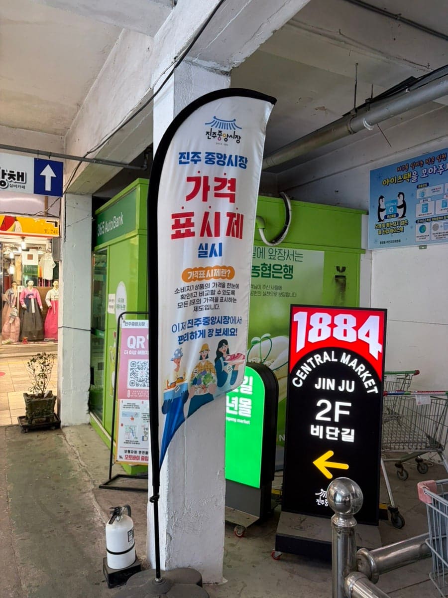 1884년을 새긴 진주중앙시장 표지판과, 상인회가 있는 3층과 청년몰 비단길이 있는 2층을 가리키는 안내판이다. 가격표시제를 알리는 현수막도 함께 걸려 있다.