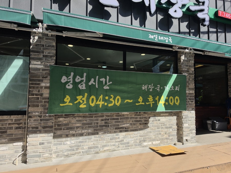 rymon은 햇빛 환한 낮 시간 영업시간 현수막이 걸린 가게 외관과, 기와 지붕 모양으로 꾸민 나무 간판 아래 조리대에서 음식을 준비하는 모습을 찍었습니다.