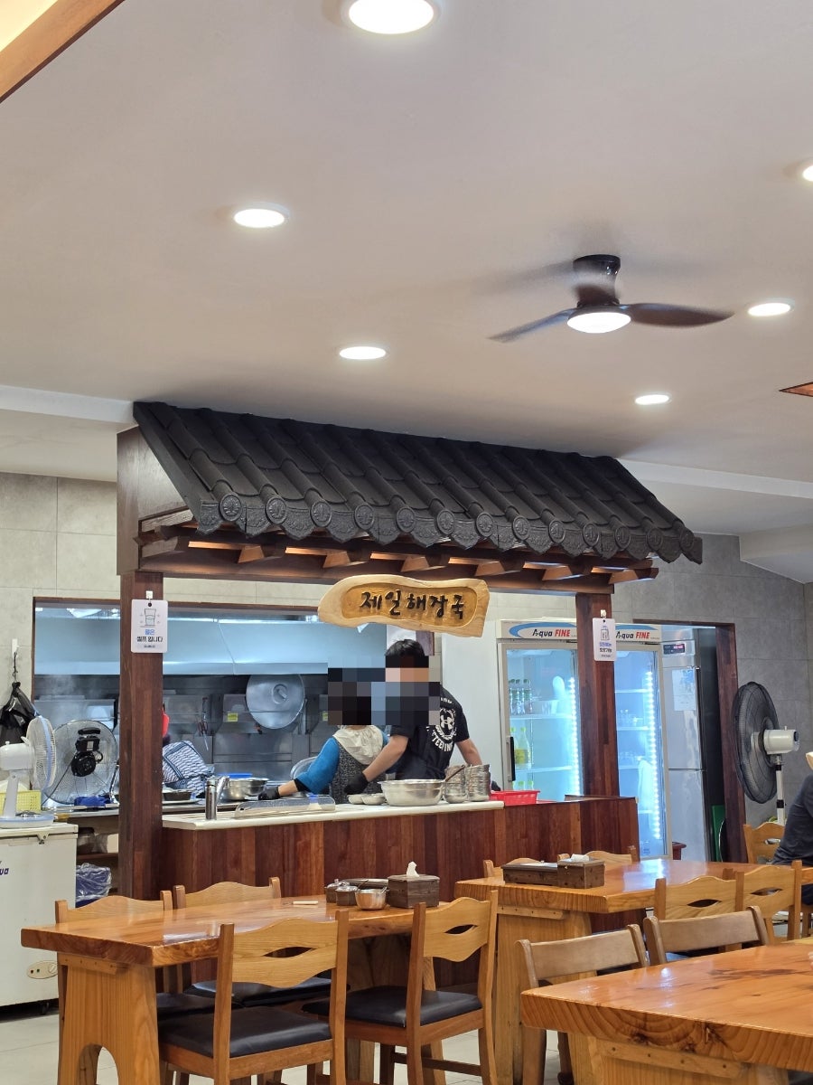 rymon은 햇빛 환한 낮 시간 영업시간 현수막이 걸린 가게 외관과, 기와 지붕 모양으로 꾸민 나무 간판 아래 조리대에서 음식을 준비하는 모습을 찍었습니다.