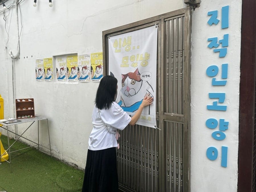 철문 앞에 서서 포스터를 붙이던 손님의 시점입니다. 하늘색 글씨로 크게 쓴 간판 옆, 고양이가 그려진 종이가 문에 붙어 있습니다.