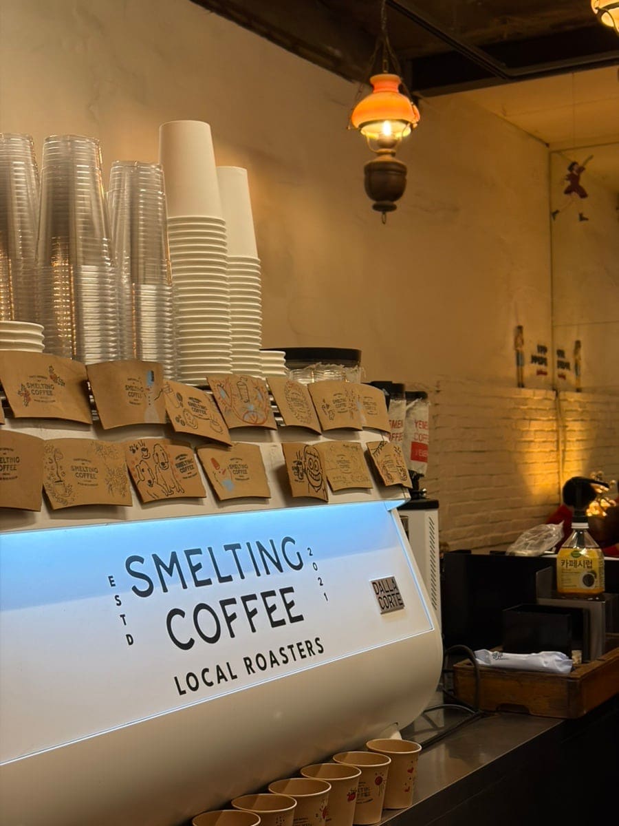 컵 홀더마다 손그림이 그려져 있고, 벽엔 'SMELTING COFFEE LOCAL ROASTERS' 글자가 커다랗게 적혀 있습니다. 평일 오전 여덟 시부터 문을 열고,