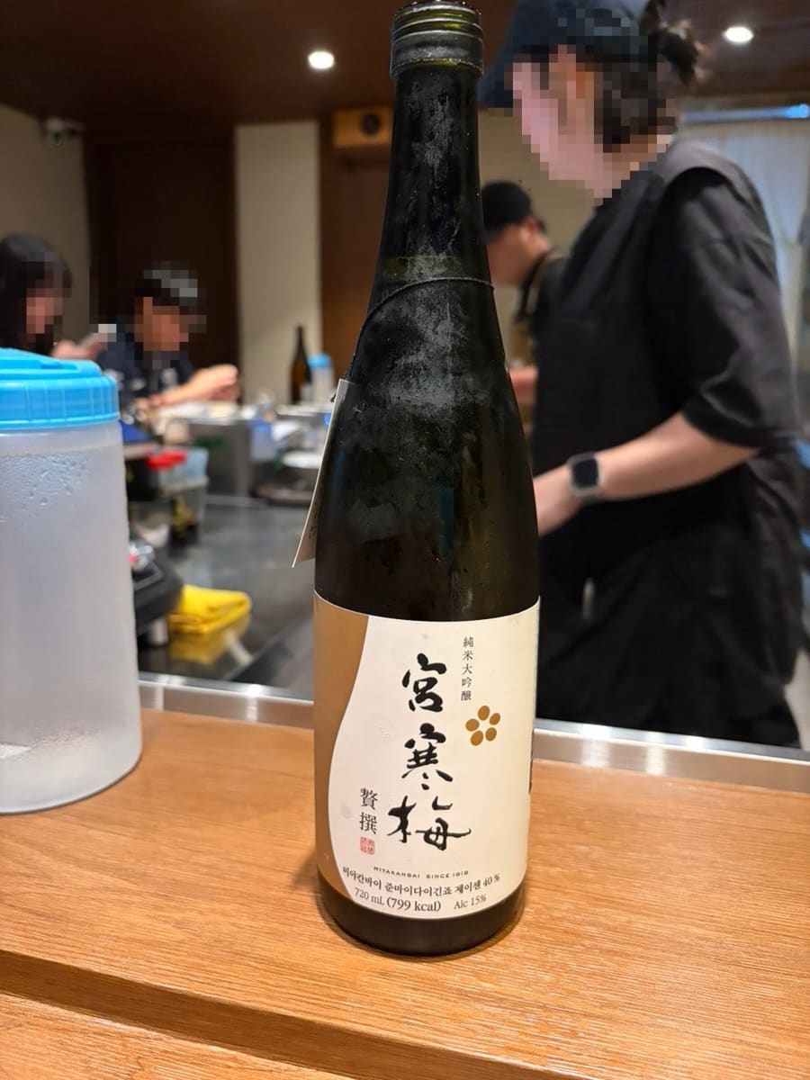오마카세로 개편된 뒤 데이트로 찾은 손님의 기록입니다. 오픈 주방을 등지고 놓인 사케와, 김가루·참깨·파를 수북이 얹은 들기름 비빔소바가 놓여 있습니다.