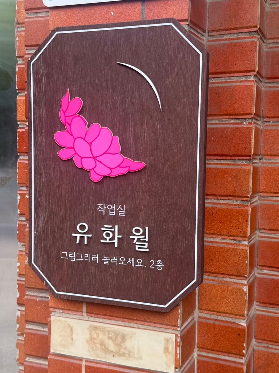 com108은 문 안으로 들어서자마자 감성적인 내부 공간이 펼쳐졌다고 적었습니다. 벽돌 벽에 붙은 갈색 이름판에는 작업실 유화월이라는 글씨와 분홍 꽃 모양이 있고,