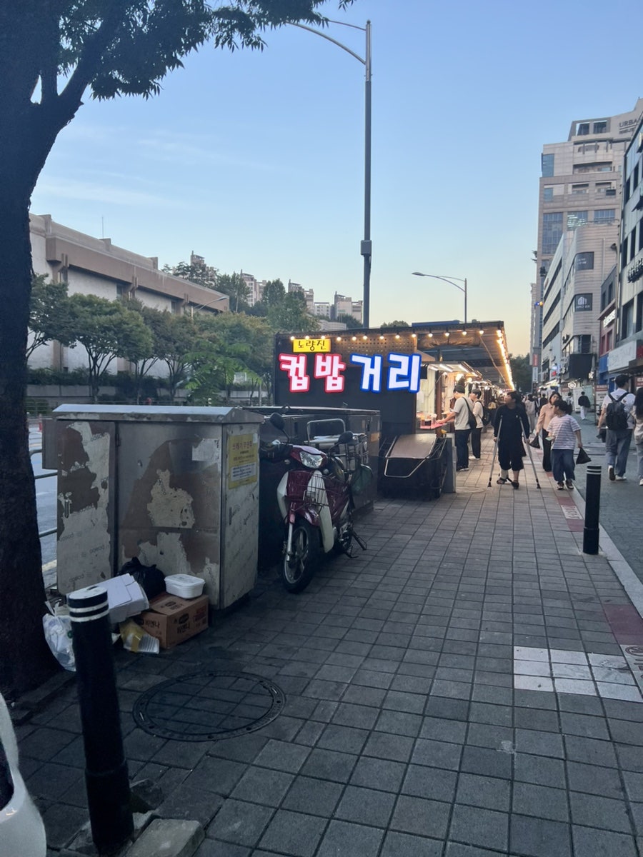 kar7519가 담은 노량진 컵밥거리 입구입니다. 저녁빛 속에 네온 간판이 켜지고, 지붕을 덮은 노점들이 길게 줄지어 있습니다.
