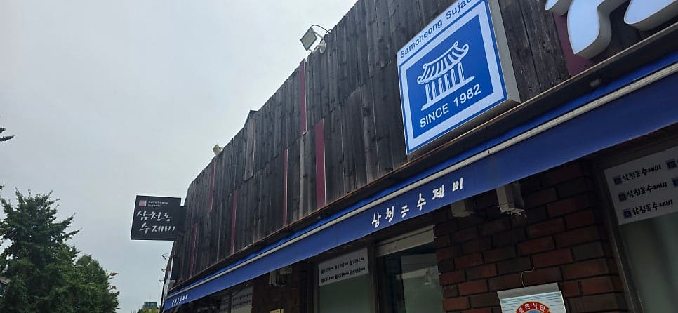 ehwalo82는 삼청동주민센터 맞은편에 자리한 소담한 가게라고 적었습니다. 사진에는 어두운 나무 벽에 파란 차양을 두른 낮은 건물 전체와,
