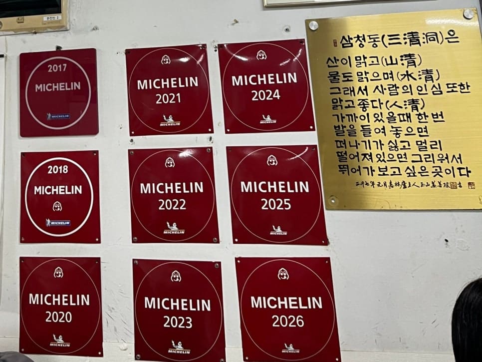 hongkeytongkey가 찍은 벽에는 붉은 미쉐린 패널이 여러 줄로 붙어 있고, 옆에는 삼청동이라는 이름을 풀이한 금색 현판이 걸려 있습니다.