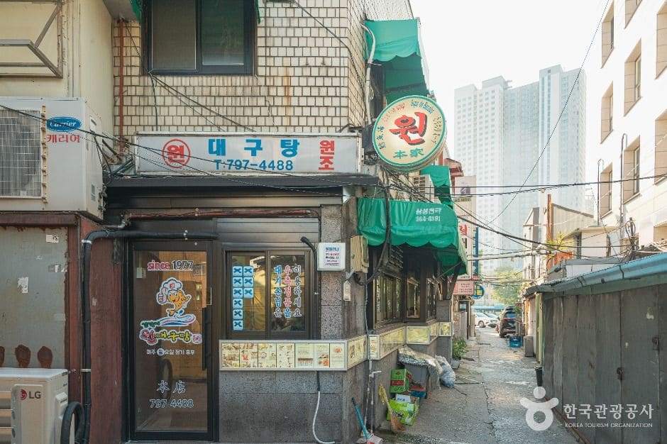 '원대구탕' 간판이 붙은 노포 앞 골목입니다. 초록 차양과 빨간 글씨 간판 사이로 좁은 골목이 이어지고, 골목 끝으로 용산의 고층 아파트가 대비되어 보입니다.