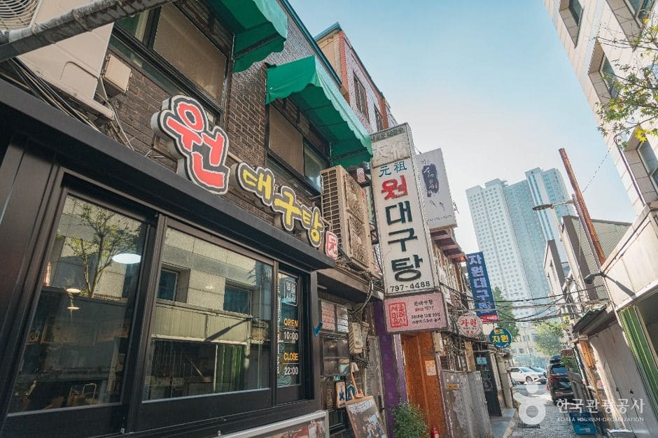 '원대구탕' 간판이 붙은 노포 앞 골목입니다. 초록 차양과 빨간 글씨 간판 사이로 좁은 골목이 이어지고, 골목 끝으로 용산의 고층 아파트가 대비되어 보입니다.