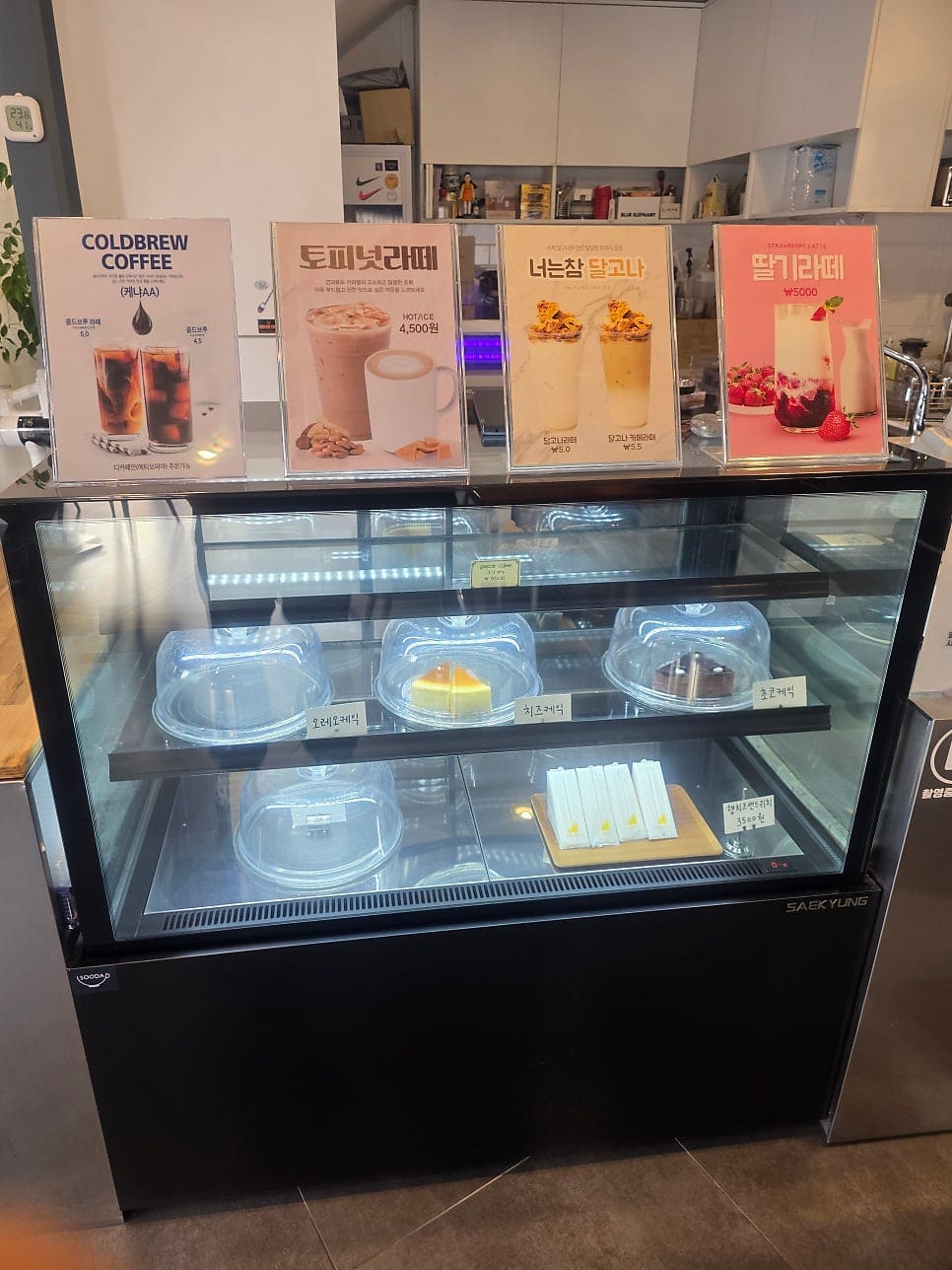 창가 테이블에 놓인 작은 화분과, 4인 이상만 앉아 달라는 손글씨 안내판입니다. 다른 사진에는 케이크 냉장고 위로 콜드브루와 토피넛라떼,