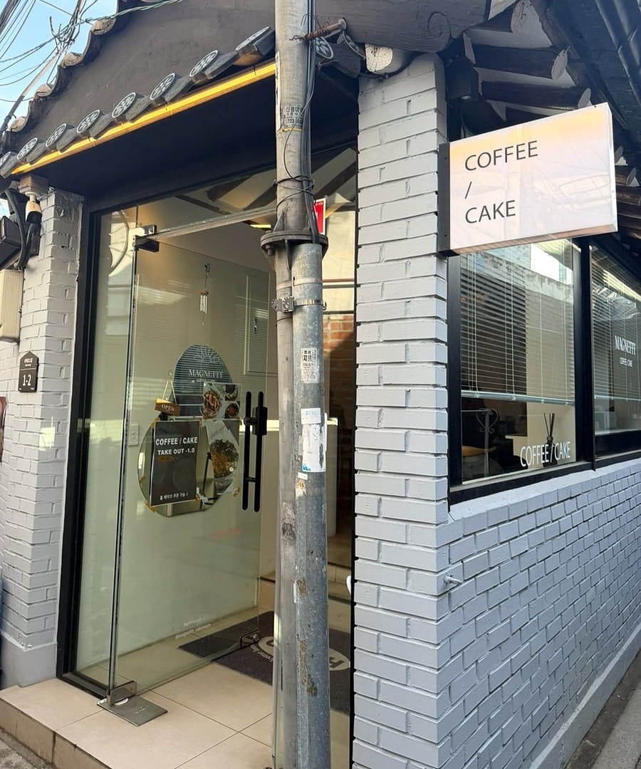 aaaxsss는 기와지붕 아래 하얀 벽돌 외관부터 담았습니다. 유리문에 붙은 동그란 로고와 'COFFEE / CAKE' 간판이 골목 모퉁이에서도 눈에 띕니다.