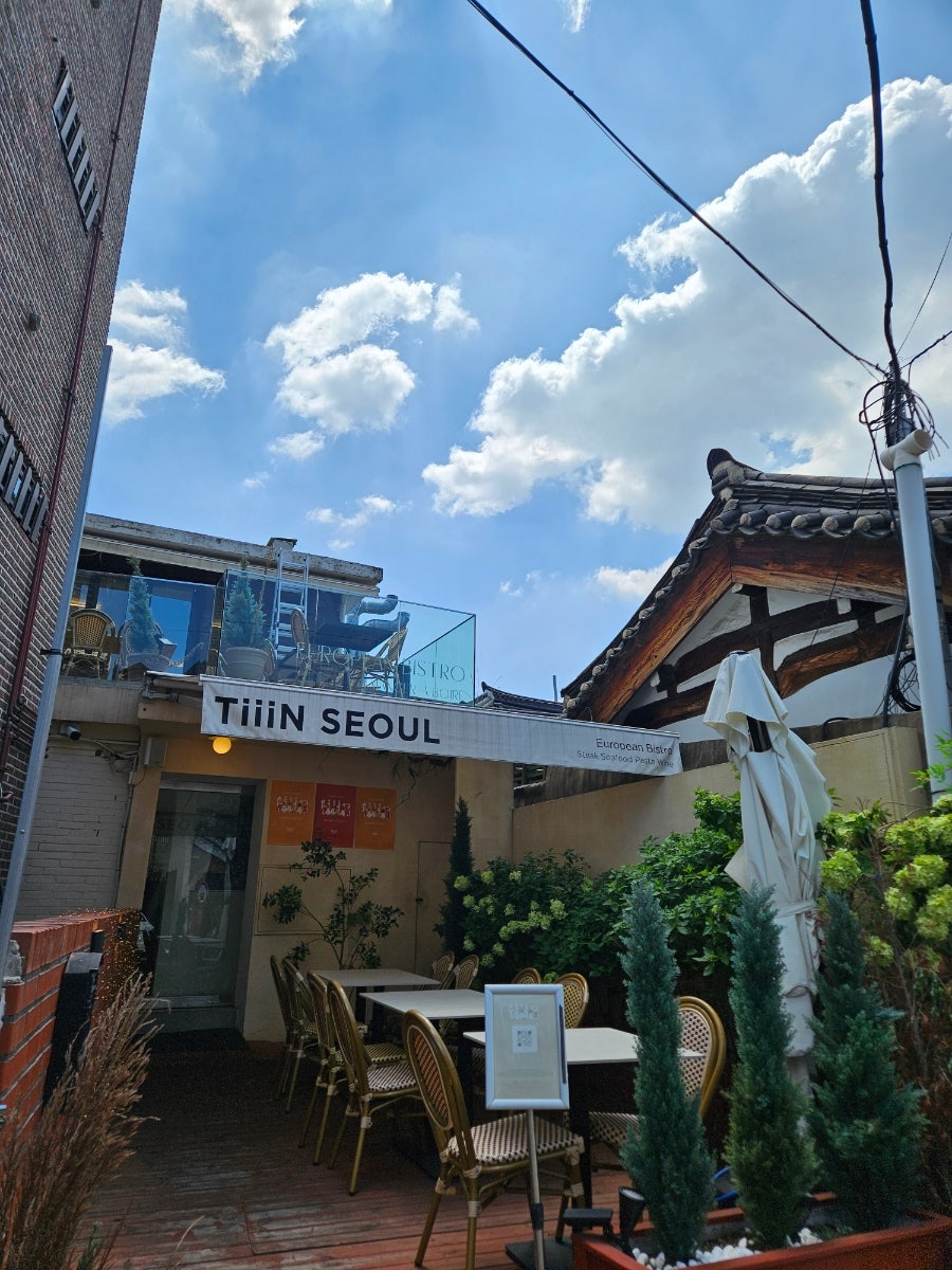 lovely_atelier는 2층 창가 테이블 너머로 펼쳐진 기와지붕 풍경과, 'TiiiN SEOUL' 간판이 걸린 테라스 입구를 찍었습니다.
