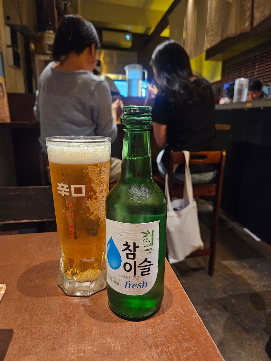 eloise02는 좁은 주방에서 요리하는 사람의 뒷모습과, 생맥주와 참이슬이 놓인 어둑한 테이블을 함께 찍었습니다.