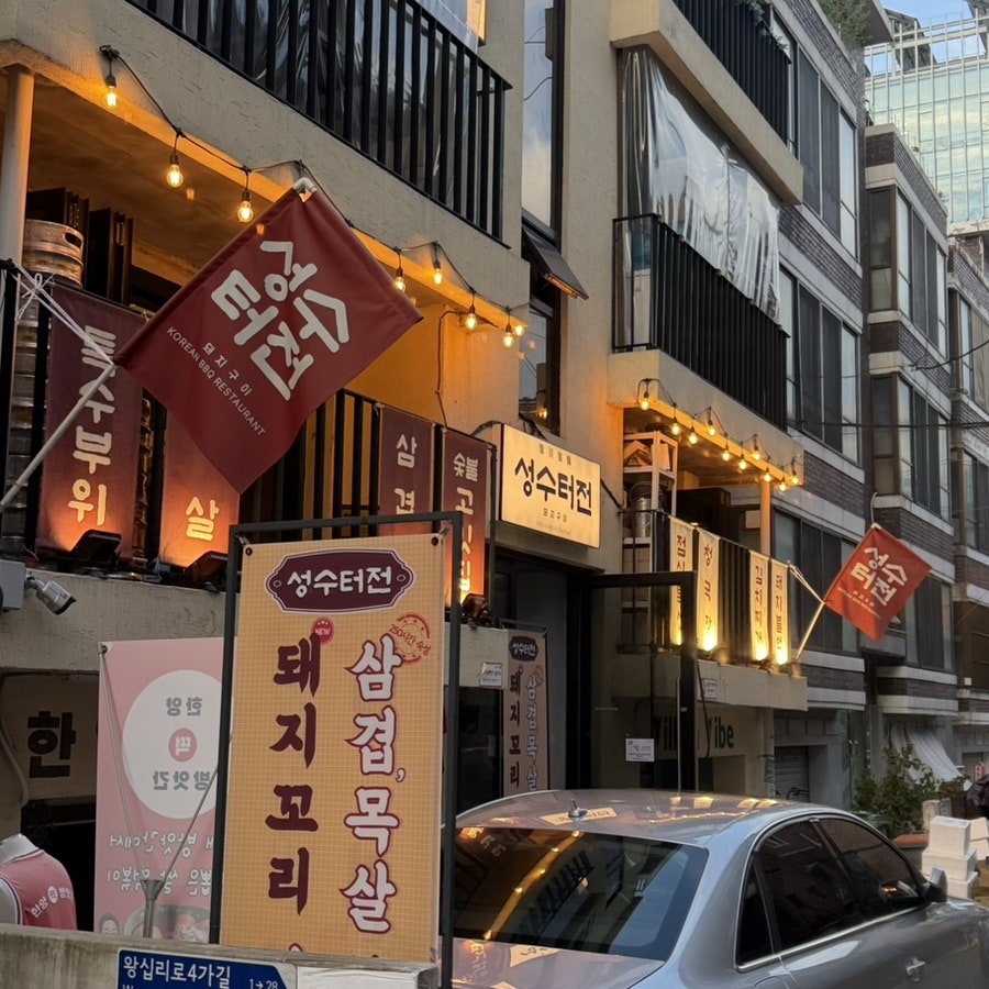 cincoj가 찍은 저녁의 건물 외관입니다. 붉은 깃발 여러 개가 나란히 걸려 있고, 바닥에 '왕십리로4가길'이라는 도로명 표지판도 함께 찍혔습니다.