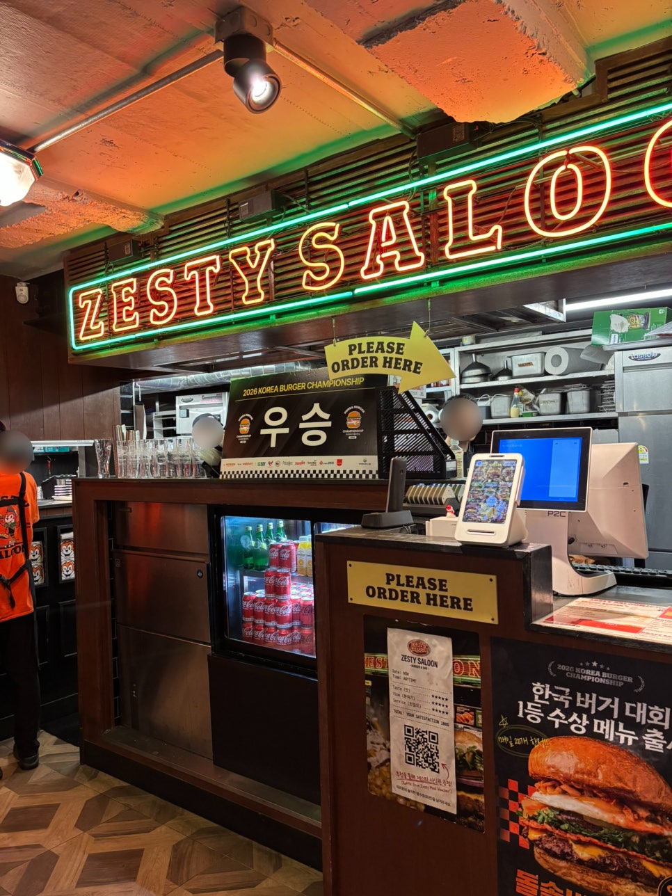네온사인 'ZESTY SALOON' 아래 '2026 KOREA BURGER CHAMPIONSHIP 우승' 트로피와 안내판이 카운터에 놓여 있습니다.