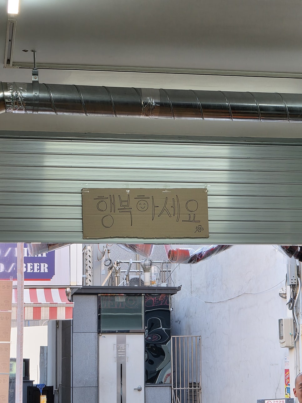 입구 셔터 위에 손글씨로 적힌 행복하세요라는 문구를 찍었습니다. 그 너머로 맞은편 건물과 골목이 함께 보여서, 가게 하나가 아니라 그 앞 골목까지 눈에 들어오는 사진입니다.
