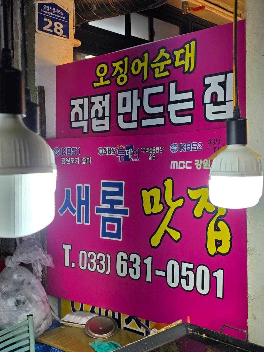 among0319가 찍은 가게 입구에는 '새롬맛집 수제오징어순대'라는 간판이 걸려 있고, 그 아래 '오징어순대 1팩 28,000원'이라는 손글씨 안내판이 놓여 있습니다.