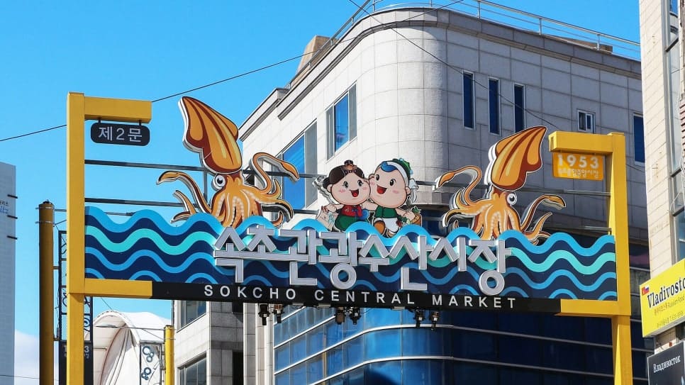 xeongdo_가 올려다보고 찍은 제2문 출입구에는 파란 하늘을 배경으로 'SOKCHO CENTRAL MARKET'과 함께 '1953'이라는 숫자가 노란 기둥에 박혀 있습니다.