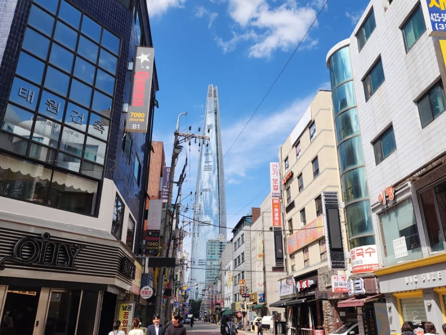 herutopia가 촬영한 송리단길 초입 거리 풍경입니다. 롯데타워가 멀리 보이는 이 대로변에서 상가 간판이 빼곡히 늘어선 골목의 규모를 짐작할 수 있습니다.