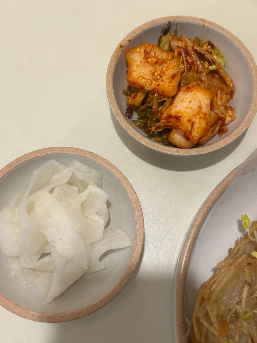 yummyzip_이 찍은 매장 안쪽과 기본 찬입니다. 벽에는 들기름막국수를 담은 사진 포스터가 액자로 걸려 있고, 나무 의자와 정갈하게 정돈된 수저통이 테이블마다 놓여 있습니다.