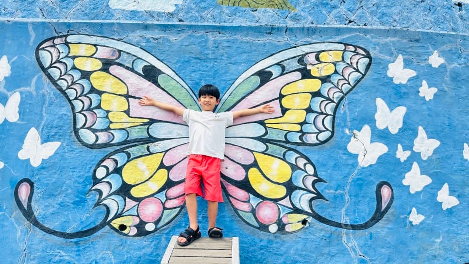 깨진 타일 조각으로 채운 고양이 모자이크 벽화를 들여다보는 아이들과, 나비 날개 벽화 앞에서 팔을 벌리고 선 아이입니다. 포토존마다 걸음이 자연스럽게 멈춘다.