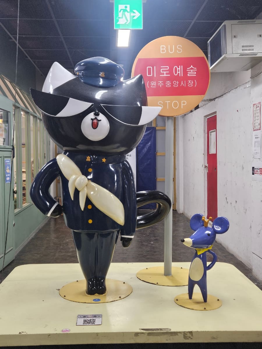 원주중앙시장 2층 입구에 들어서면 '미로예술(원주중앙시장)' 버스 정류장 표지판과 순찰모를 쓴 까만 고양이 마스코트 조형물이 먼저 손님을 맞는다고 konari33은 적었다.