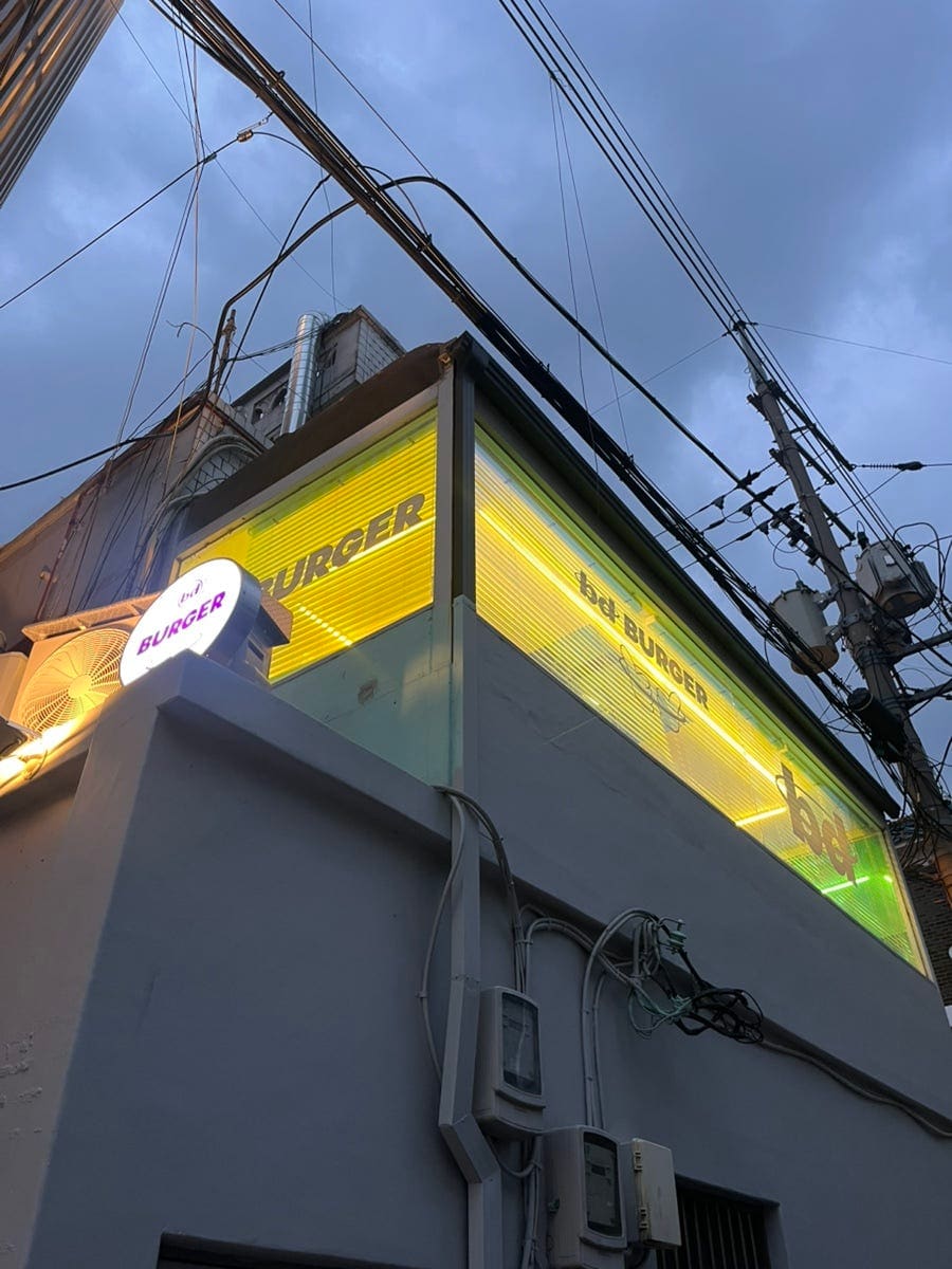 silvinha가 촬영한 bd버거의 다른 얼굴입니다. 노란 조명이 들어온 간판이 담긴 밤거리 사진과, 계단 벽면을 가득 채운 포스터·굿즈 사진을 함께 남겼습니다.