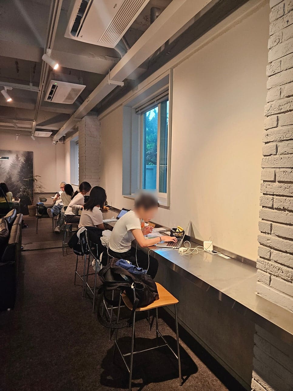 넓은 창과 높은 천장이 개방감 있는 녹녹의 공간을 더욱 돋보이게 합니다. 블로거는 모던한 인테리어와 식물로 가득한 이곳에서 책을 읽고 음료를 마시는 사람들의 모습을 담으며,