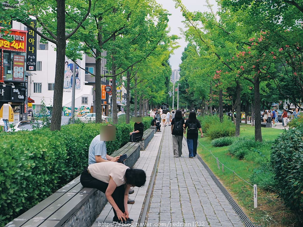 가로수 그늘이 길게 드리운 산책로를 두 사람이 나란히 걷고 있습니다. 벤치에 앉아 신발끈을 고쳐 매거나 잠시 머무는 사람들도 같은 그늘 아래 섞여 있습니다.