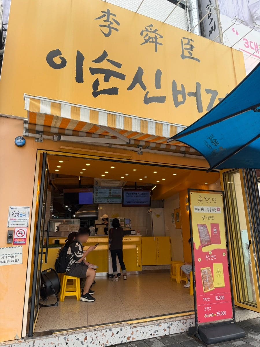 boram95_가 담은 이순신광장 골목의 이순신버거입니다. 노란 간판 아래 자리를 잡은 사람들과, 방금 받아든 노란 포장 봉투가 여행의 간식 시간을 보여줍니다.