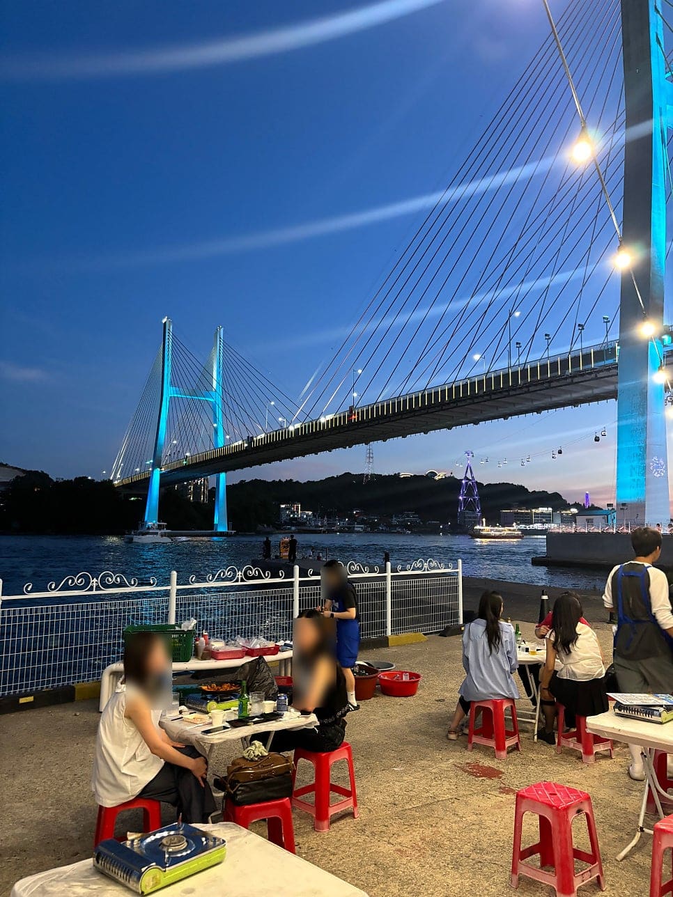 eunmi2110이 담은 테라스와 야장 자리에서 본 밤바다입니다. 파란 조명을 받은 거북선대교와 케이블카 곤돌라가 지나가고, 사람들이 빨간 플라스틱 의자에 둘러앉아 있습니다.