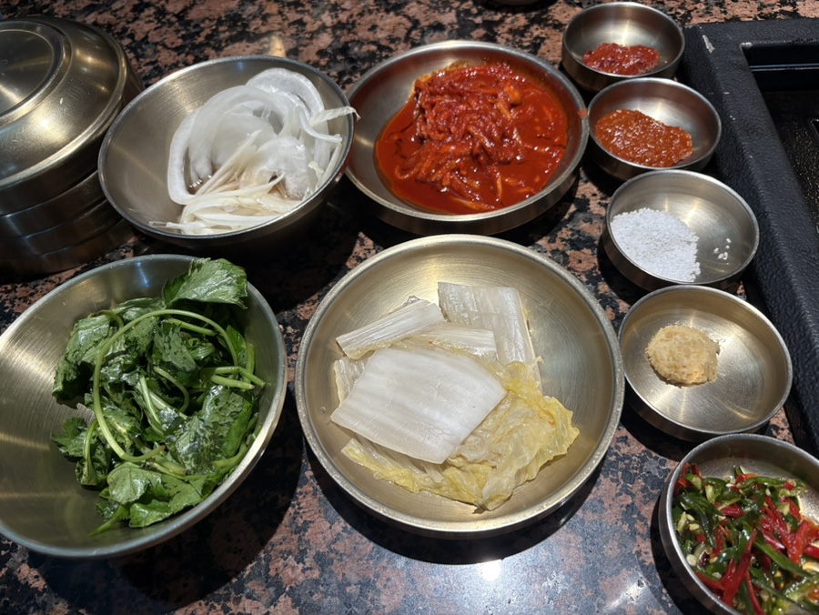 og_log가 담은 장면입니다. 앞치마를 두른 직원이 그릴 앞에서 고기를 손질하고, 테이블 위에는 미나리와 묵은지, 양념장, 소금, 마늘까지 곁들임 그릇이 가득합니다.